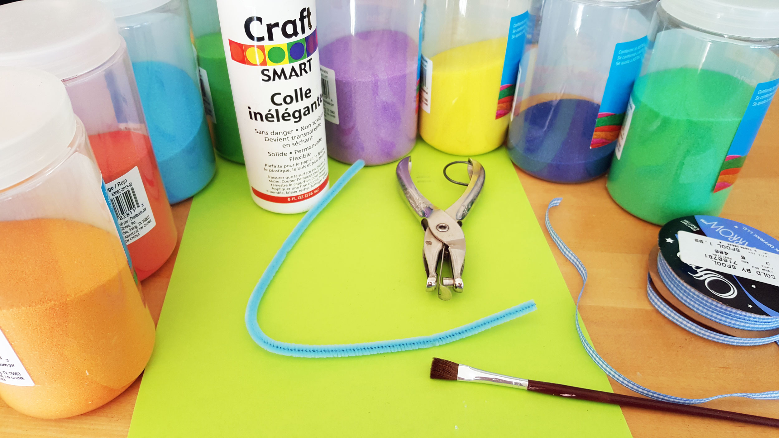 Sand art supplies needed for your DIY ornament: Cardstock paper, scissors, colored sand, glue, paintbrush, hole puncher, pipe cleaners, and ribbon | OrnamentShop.com