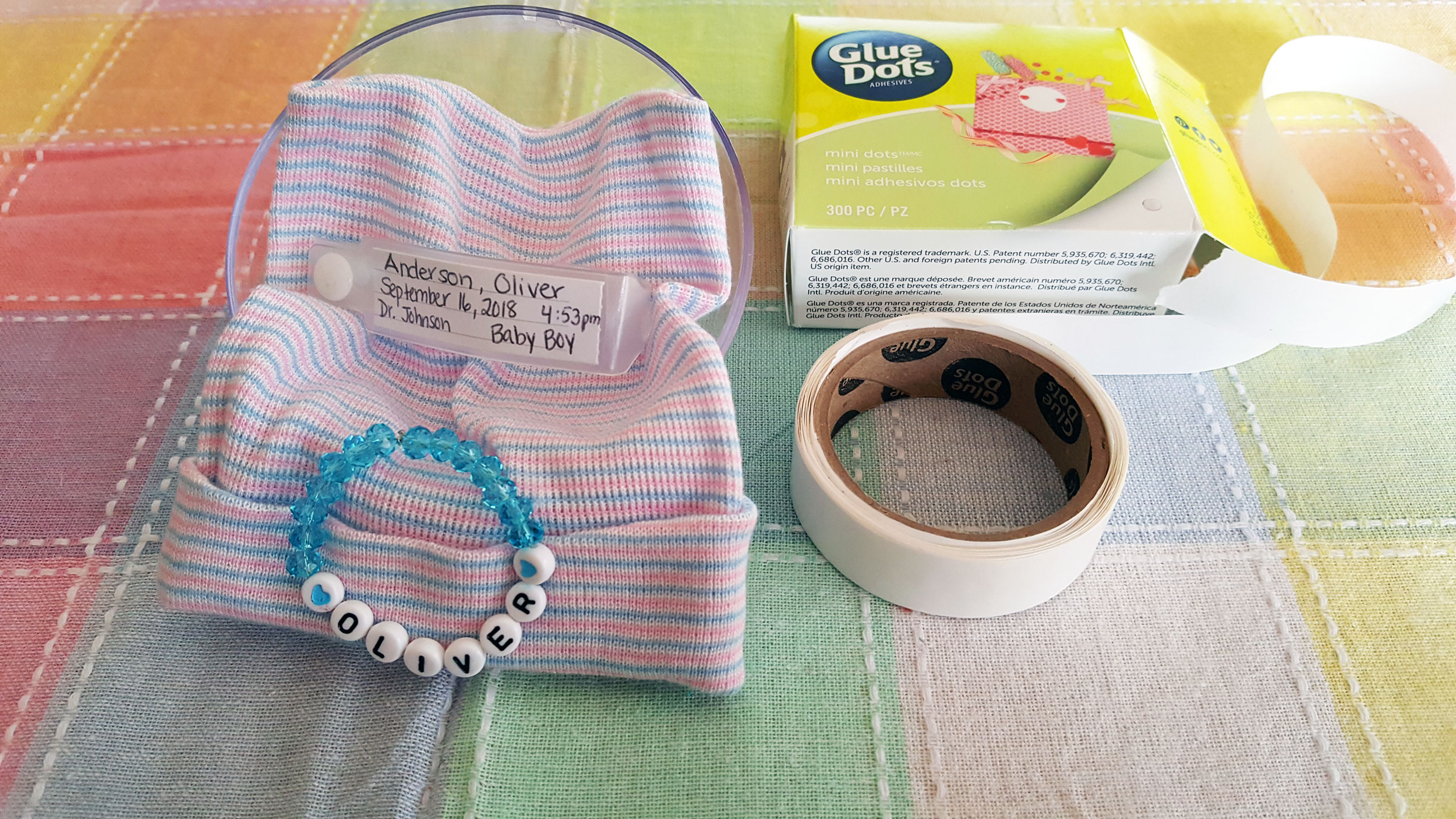 Newborn's name in beads (bracelet), newborn hat and delivery bracelet, masking tape, and glue dots on table. | OrnamentShop.com